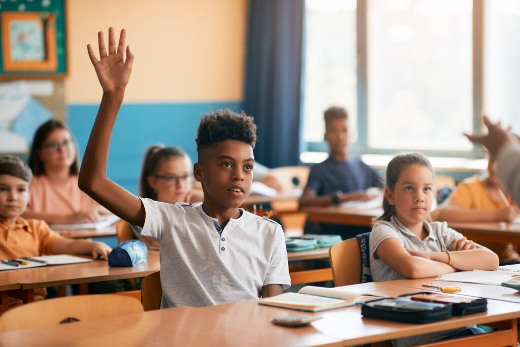 Black elementary student raising his hand to answer teacher's question in the classroom.